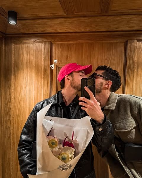 two boys kissing behind a phone, a bouquet in hand